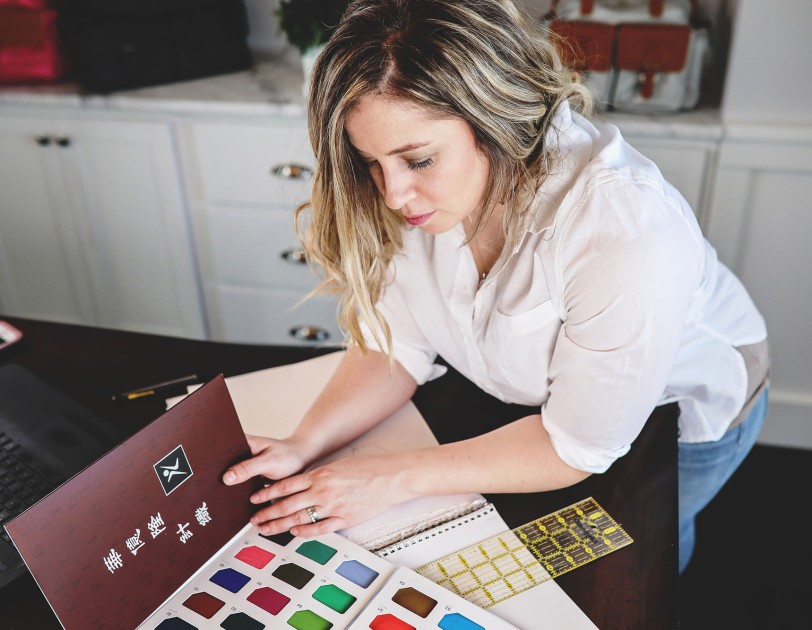 A woman wearing a white button down and blue jeans holds a fabric swatch book, examining the contents of the book. She leans over a desktop, which also holds her computer, notebook, and other office supplies.